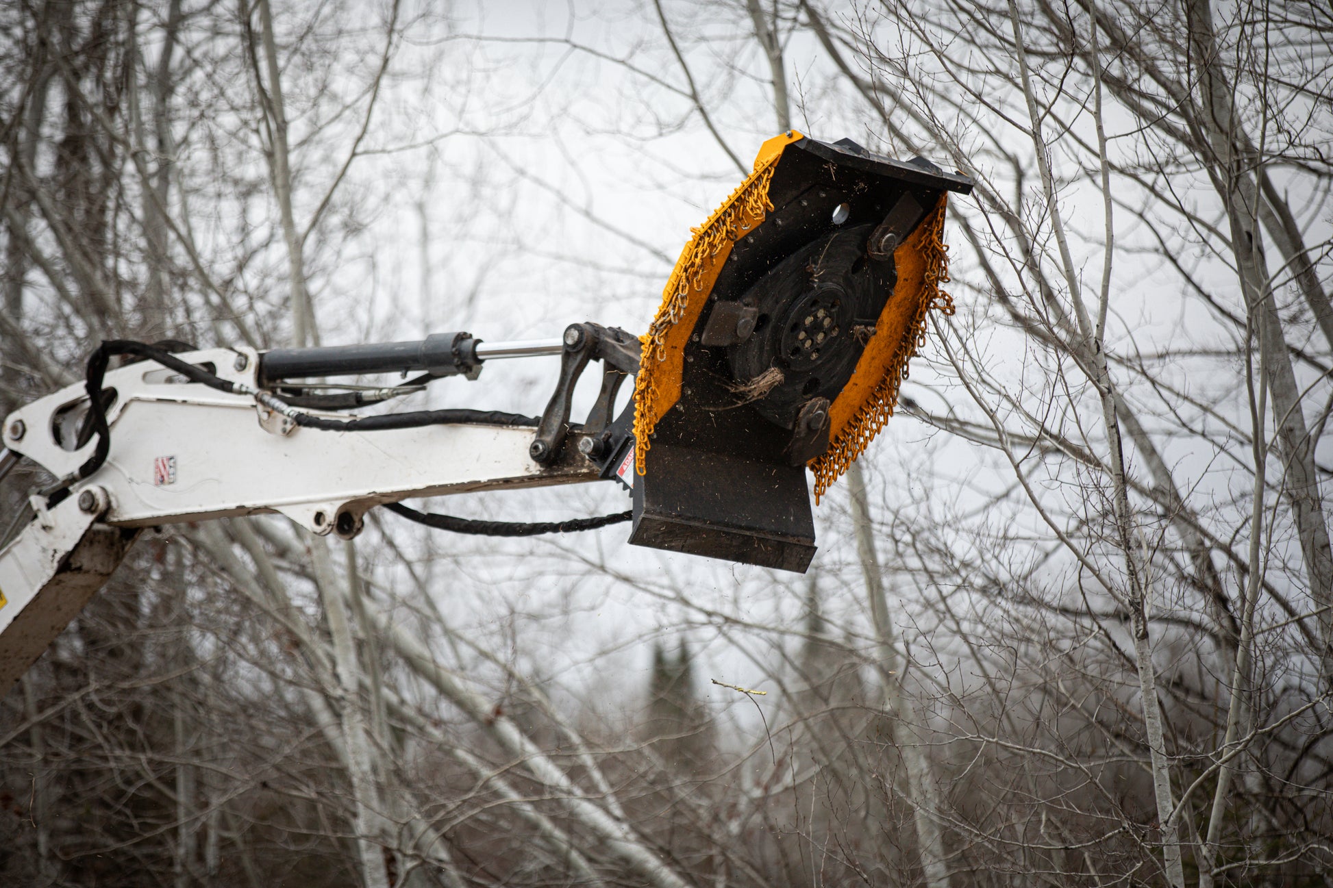 Excavator arm with a mower attachment against a forest background.