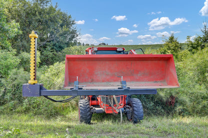 Front view of a red tractor with large red bucket with the Bucket Mount Limb Cutter attachment extended beside brush.