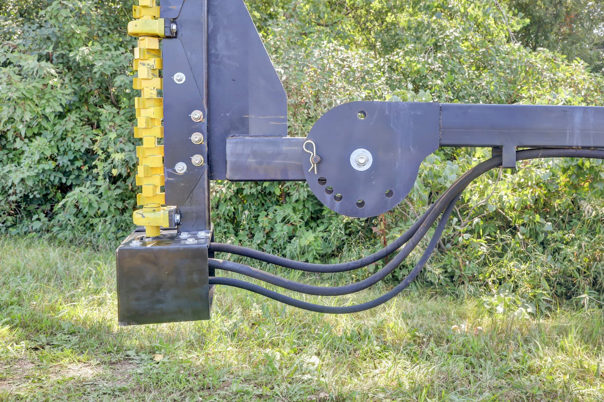 Close-up of the Quick Attach Compact Limb Cutter’s yellow cutting rotor, adjustable black mounting arm and hydraulic hoses beside dense brush.