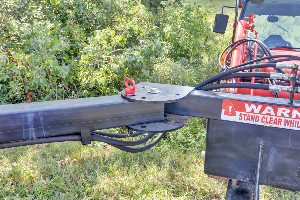 Close-up of the Compact Limb Cutter’s black mounting arm and hydraulic hoses attached to a red tractor beside dense brush.