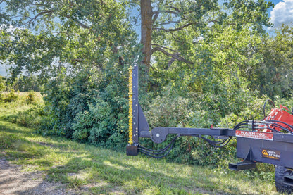 Quick Attach Compact Limb Cutter mounted on a red tractor beside a tree and dense brush, with its yellow cutting rotor upright.