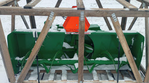 A green snowblower attached to a wooden structure, set in a snowy environment.