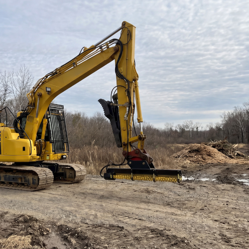 A yellow excavator with a large black and yellow brush cutter attachment working on a muddy construction site under a cloudy sky.