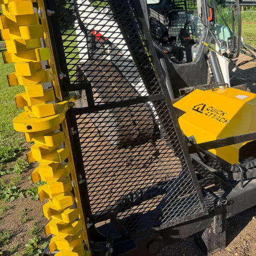 Close-up of brush cutter attachment with a black metal mesh gate and yellow cutting teeth, mounted to a yellow skid steer.