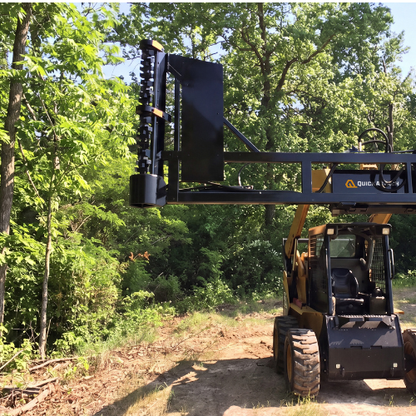 A yellow and black skid steer with a large black brush cutting attachment arm raised overhead, photographed outdoors on a dirt path surrounded by green trees and brush.