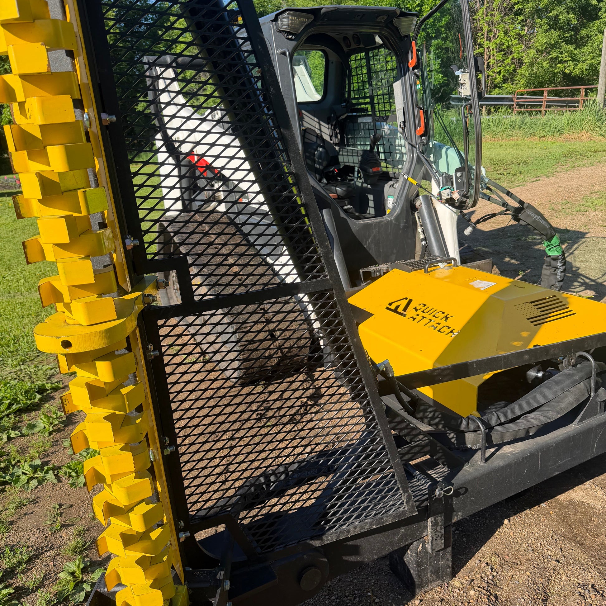 A close-up outdoor shot of a yellow and black limb cutter attachment, showing a metal mesh guard, yellow teeth or tines, and part of a cab in a dirt and grass field.
