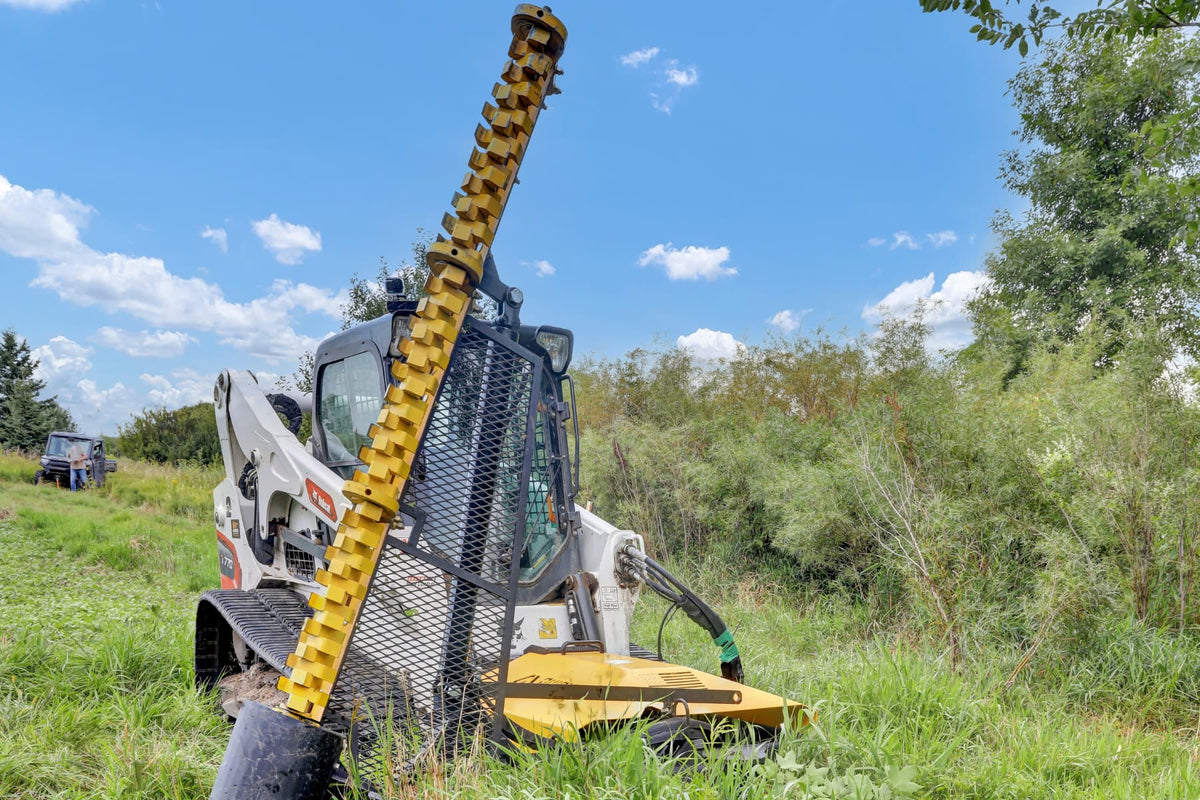 Front view of the Limb Cutter 6 raised beside a skid steer at the edge of a field.