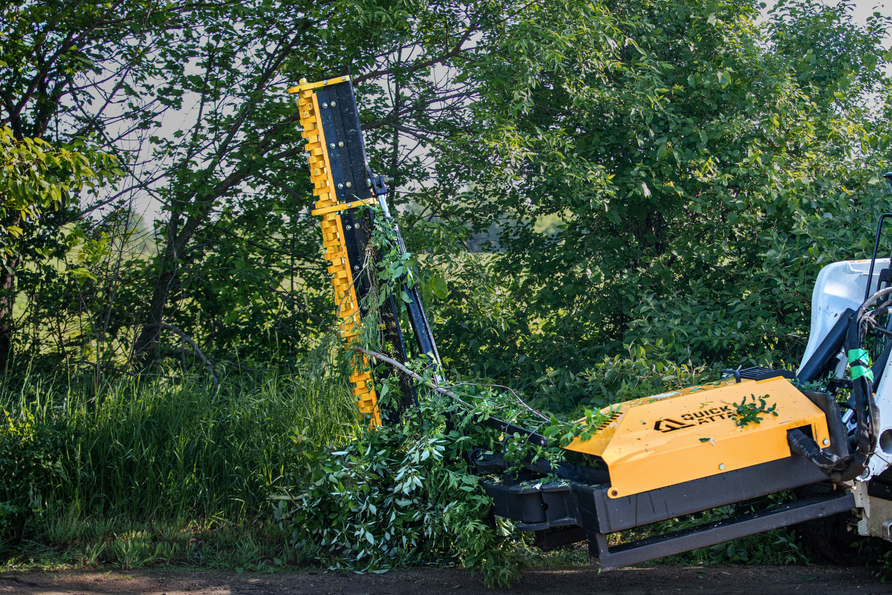 A yellow and black stump grinder attachment mounted on a white vehicle, shown clearing green tree stumps and brush outdoors in a wooded area.