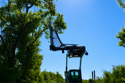A tractor-mounted tree trimmer or pruner attachment is shown in a real outdoor setting with green trees and a clear blue sky. A person is visible inside the tractor cab, so this is classified as Lifestyle rather than Product/Packaging.