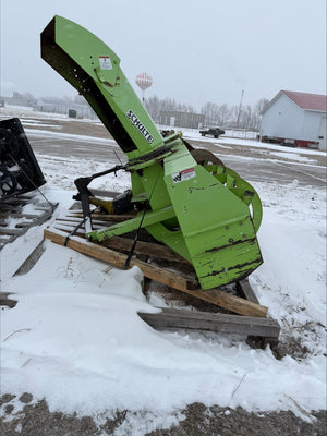 Green snowblower on a snowy ground with a building and vehicle in the background.