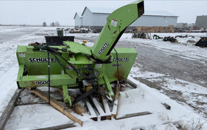 Green Schulte snow blower on a snowy ground with buildings in the background